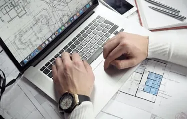 Hands typing at a laptop on a desk strewn with printed floor plans, a phone and drafting pens.