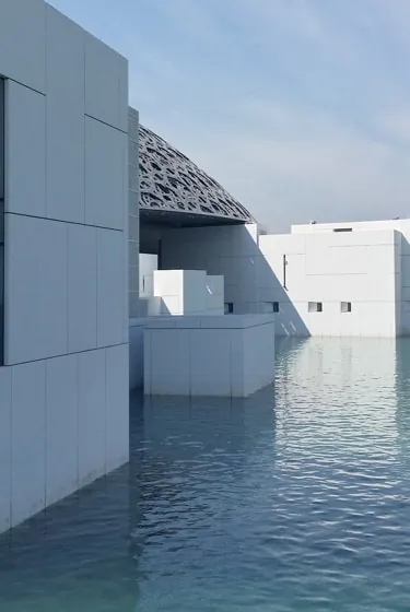 White cubic museum pavilions standing in still, shallow water, a perforated steel dome rising behind them.